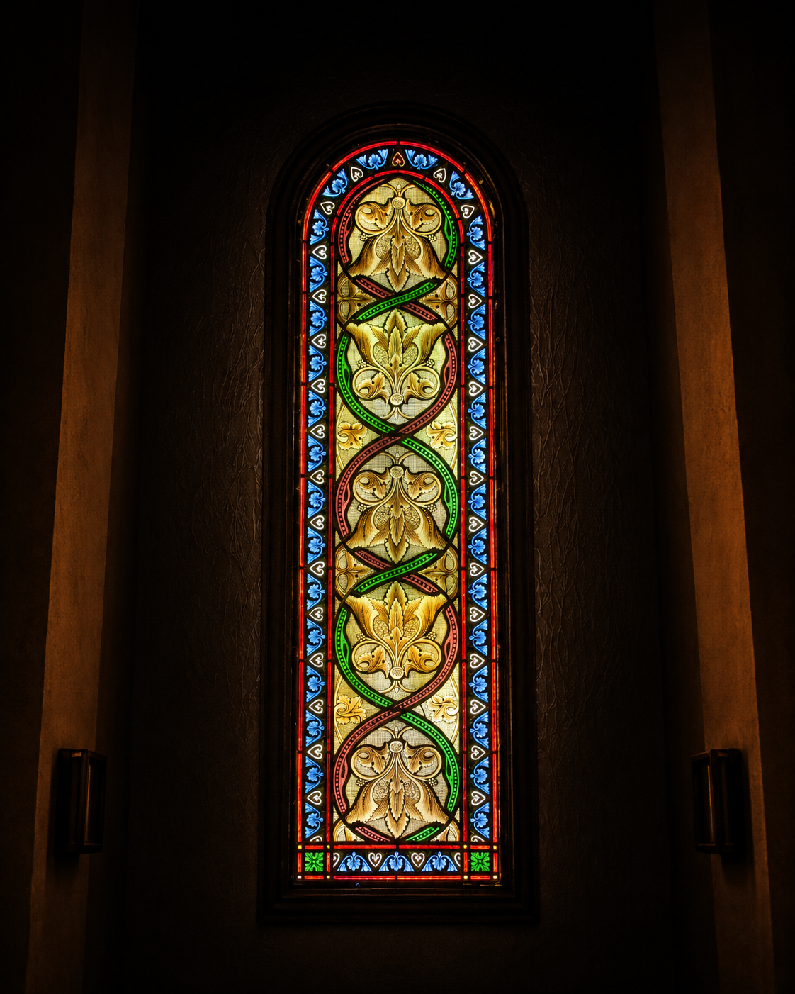 Vitrail traditionnel vertical orné de motifs floraux dorés, bordures bleues, rouges et vertes, dans une chapelle